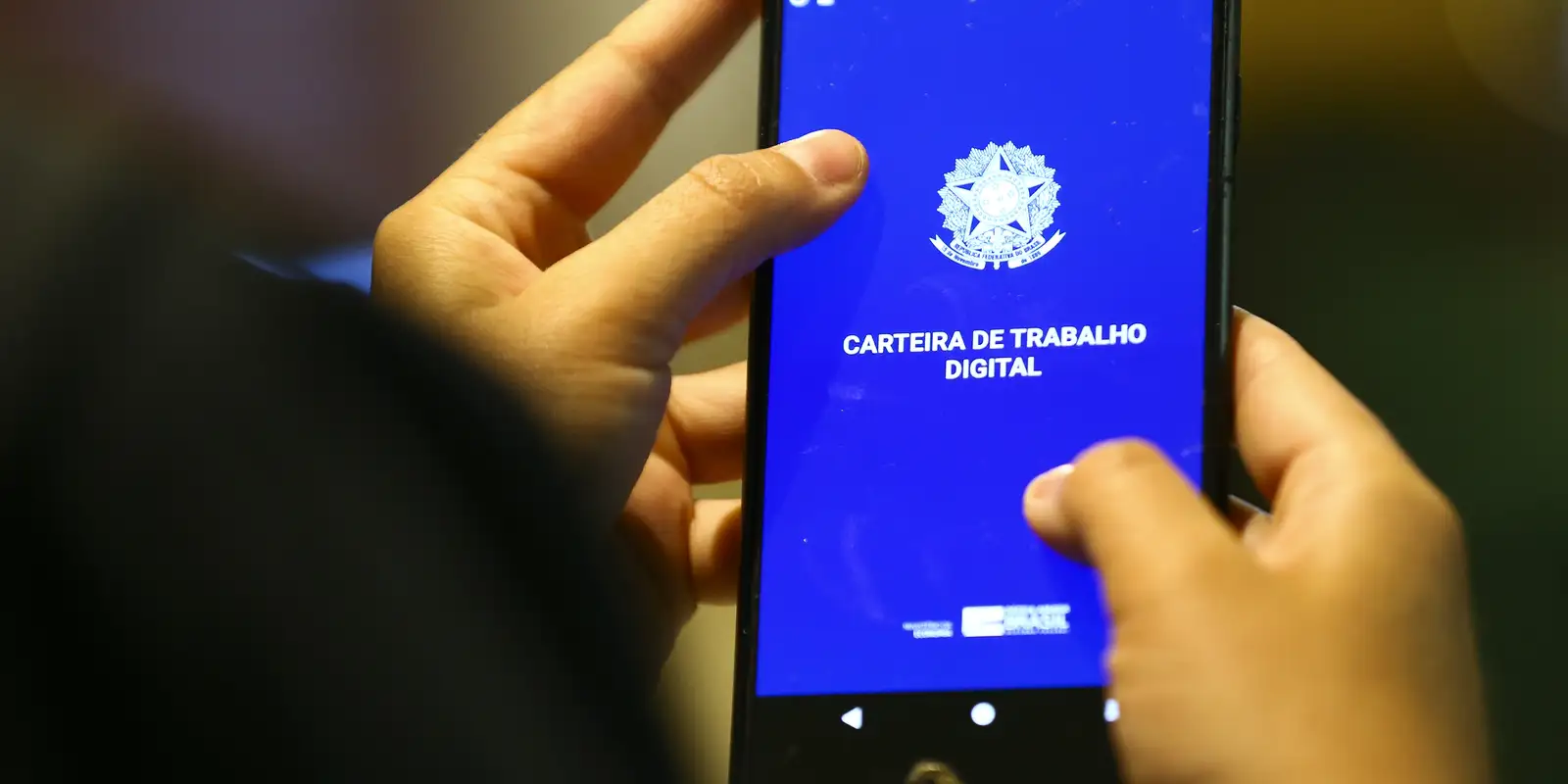 Governo libera parcelas extras do Seguro-Desemprego para cerca de 13,5 mil pessoas afetadas pelas chuvas em Minas Gerais 2 Mãos segurando celular com tela da Carteira de Trabalho Digital azul