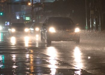 Carro com faróis acesos trafegando por uma rua urbana alagada durante uma tempestade noturna, com reflexos das luzes na água e chuva intensa.