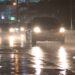 Carro com faróis acesos trafegando por uma rua urbana alagada durante uma tempestade noturna, com reflexos das luzes na água e chuva intensa.