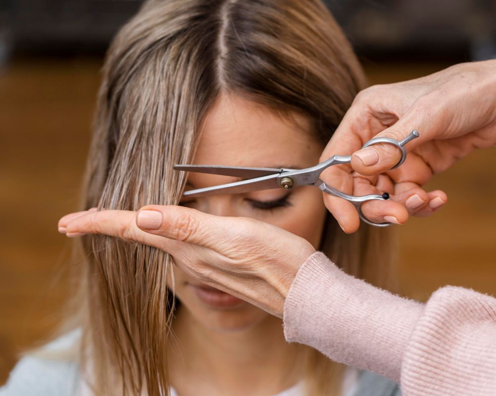 Corte de cabelo e renovação emocional: Por que muitas pessoas escolhem mudar a aparência durante períodos de crise? 2 Close de uma pessoa cortando a própria franja reta com uma tesoura prateada, com foco nas mãos e no movimento cuidadoso dos fios loiros.