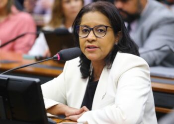 Mulher de óculos e blazer branco falando ao microfone em uma sessão da Câmara dos Deputados, representando a bancada feminina durante debates legislativos.