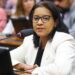 Mulher de óculos e blazer branco falando ao microfone em uma sessão da Câmara dos Deputados, representando a bancada feminina durante debates legislativos.