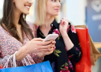 Duas mulheres sorrindo com sacolas de compras e celular nas mãos em shopping