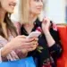 Duas mulheres sorrindo com sacolas de compras e celular nas mãos em shopping