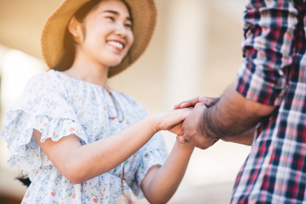 Por que pedir um pequeno favor a alguém que não gosta de você faz essa pessoa passar a te admirar 2 Close-up das mãos de um homem e uma mulher se segurando em um gesto de apoio e confiança, com a mulher sorrindo ao fundo, usando um vestido floral e chapéu de palha, em um ambiente externo ensolarado.