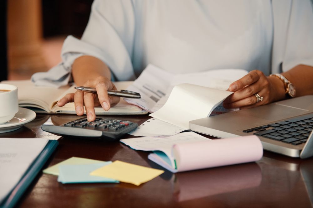 Dívidas no Banco do Brasil podem ser renegociadas com descontos de 90% até o fim do mês 2 Close-up das mãos de uma pessoa usando uma calculadora e conferindo papéis sobre uma mesa de madeira, ao lado de um notebook.
