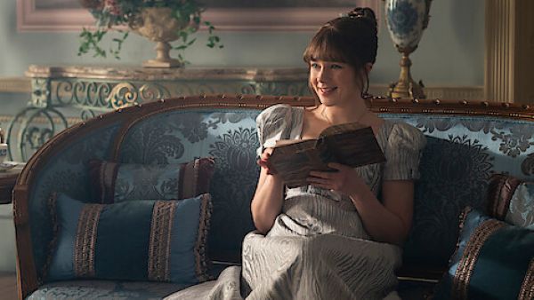 Quem será a próxima protagonista da 5ª temporada de Bridgerton? Confira! 2 Eloise Bridgerton, interpretada por Claudia Jesse, sentada em um sofá azul ornamentado, segurando um livro aberto e sorrindo enquanto olha para o lado.