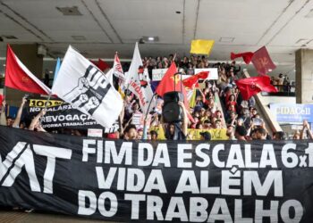 Uma multidão de manifestantes em uma escadaria segura bandeiras coloridas e uma grande faixa preta horizontal com a frase "FIM DA ESCALA 6x1 VIDA ALÉM DO TRABALHO" escrita em letras brancas garrafais.