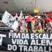 Uma multidão de manifestantes em uma escadaria segura bandeiras coloridas e uma grande faixa preta horizontal com a frase "FIM DA ESCALA 6x1 VIDA ALÉM DO TRABALHO" escrita em letras brancas garrafais.
