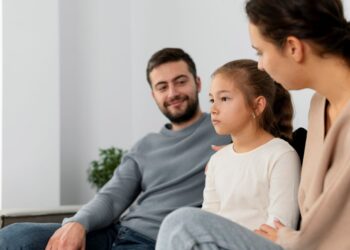 Pai e mãe sentados em um sofá conversando com a filha pequena, que mantém uma expressão pensativa e evita contato visual direto.