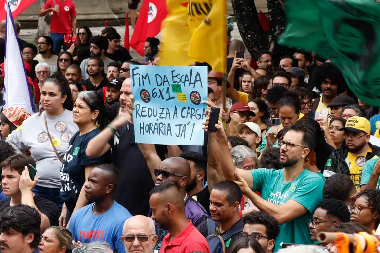 Redução da jornada semanal para 36 horas: benefícios e riscos do fim da escala 6x1 2 No centro de uma multidão em um protesto ao ar livre, um homem de óculos e camiseta verde ergue um cartaz azul claro onde se lê à mão: "FIM DA ESCALA 6x1 - REDUZA A CARGA HORÁRIA JÁ!". Ao redor, diversas pessoas participam da manifestação com bandeiras coloridas ao fundo.
