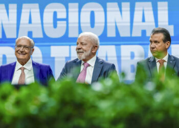 Três homens de terno sorrindo sentados à mesa com painel azul ao fundo