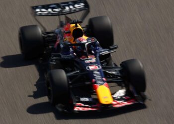 Visão aérea em ângulo oblíquo de um carro de Fórmula 1 da Red Bull Racing, com detalhes em azul marinho e bico amarelo, acelerando em uma pista cinza sob luz solar intensa.