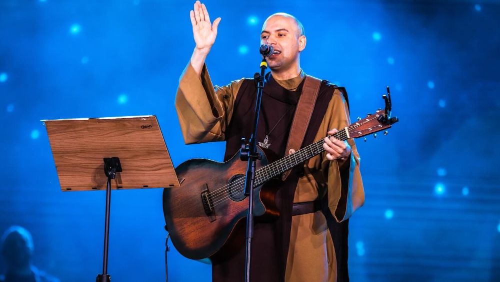 Rosário da madrugada cresce na Quaresma de 2026 e reúne fiéis antes do amanhecer em várias cidades do Brasil 2 Frei Gilson, um homem calvo vestindo hábito marrom de frade, canta ao microfone enquanto toca um violão acústico marrom em um palco com iluminação azul ao fundo.