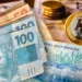 Cédulas de 100 e 50 reais ao lado de moedas de 1 real empilhadas sobre superfície