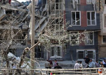 Prédio parcialmente destruído após ataque, com escombros espalhados e moradores observando os danos em área urbana no Oriente Médio.