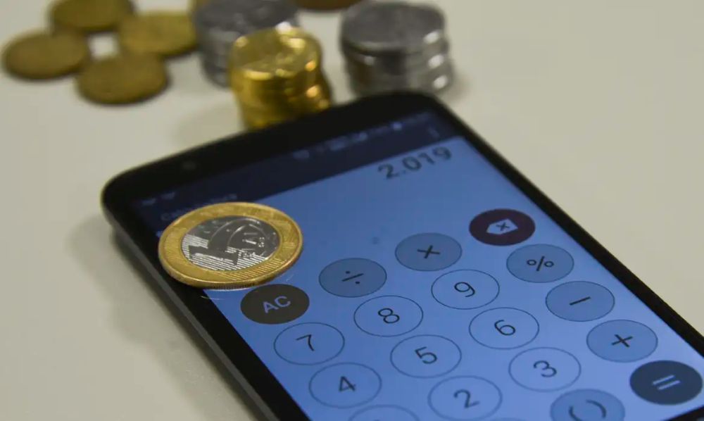 Mega-Sena sorteia R$ 60 milhões hoje (10/03); veja quanto o valor pode render 2 Moeda de um Real sobre a tela de um celular exibindo uma calculadora, com pilhas de moedas ao fundo.