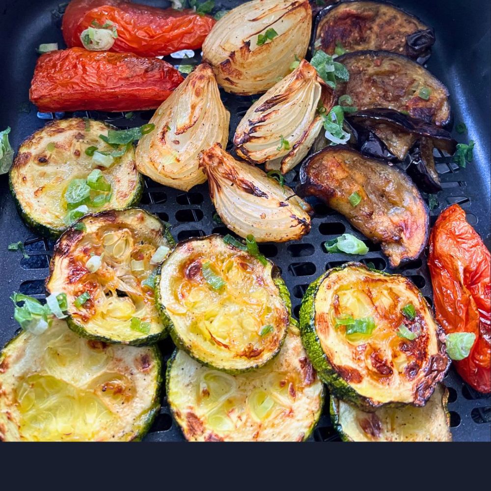 Descubra quais alimentos ficam mais saborosos na Air Fryer (melhor do que no forno tradicional)! 2 Vista de cima de uma cesta de air fryer preta cheia de rodelas de abobrinha tostadas, pedaços de cebola caramelizados, fatias de berinjela grelhadas e tomates inteiros murchos. A mistura de legumes grelhados está temperada com ervas secas e cebolinha verde picada.