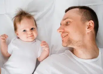 Pai sorrindo olhando para bebê deitado ao lado em cama branca