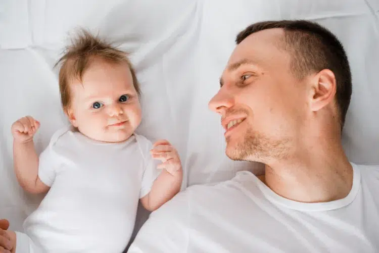 Licença-paternidade: Plenário aprova projeto que amplia o período de afastamento do trabalho 1 Pai sorrindo olhando para bebê deitado ao lado em cama branca