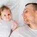Pai sorrindo olhando para bebê deitado ao lado em cama branca