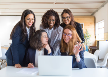 Grupo de cinco mulheres profissionais de diferentes etnias sorrindo e colaborando ao redor de um laptop em um escritório moderno.