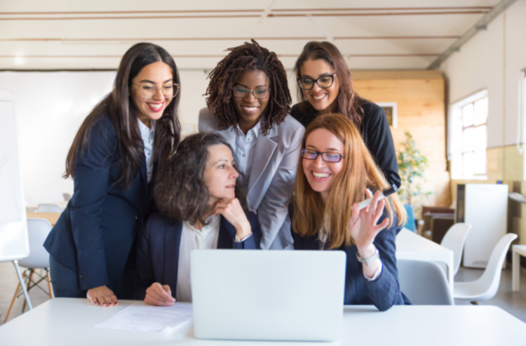 Dia das Mulheres: descubra quais profissões mais atraem o público feminino 1 Grupo de cinco mulheres profissionais de diferentes etnias sorrindo e colaborando ao redor de um laptop em um escritório moderno.