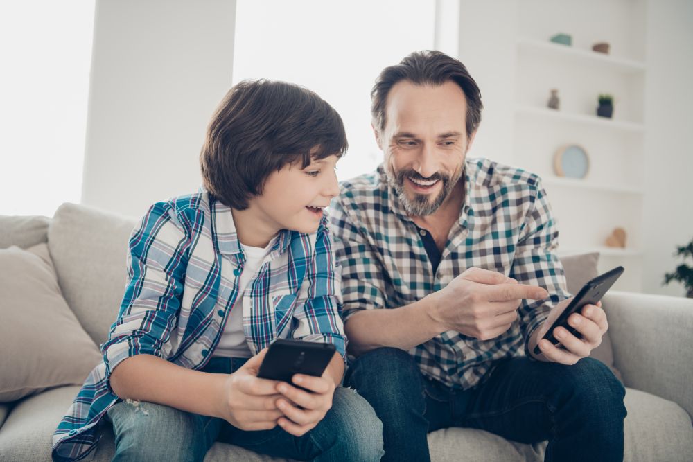 WhatsApp libera função para pais monitorarem conta dos filhos; veja como configurar 2 Homem adulto e uma criança sentados em um sofá cinza, ambos segurando smartphones. O adulto aponta para a tela do celular enquanto sorri, demonstrando como configurar ou utilizar o aplicativo de forma conjunta e segura.