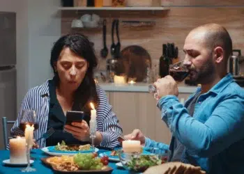 Mulher consulta celular enquanto homem bebe vinho em jantar com velas na mesa