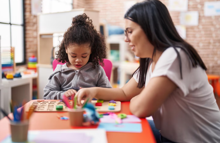 Dia das Mulheres: descubra quais profissões mais atraem o público feminino 2 Uma pedagoga jovem de cabelos escuros e óculos auxilia uma criança pequena com cabelos cacheados a montar um quebra-cabeça de letras coloridas em uma sala de aula.
