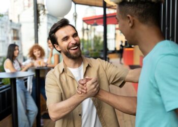 Dois homens sorridentes em um ambiente externo de confraternização, apertando as mãos de forma amigável enquanto conversam, com outras pessoas ao fundo.