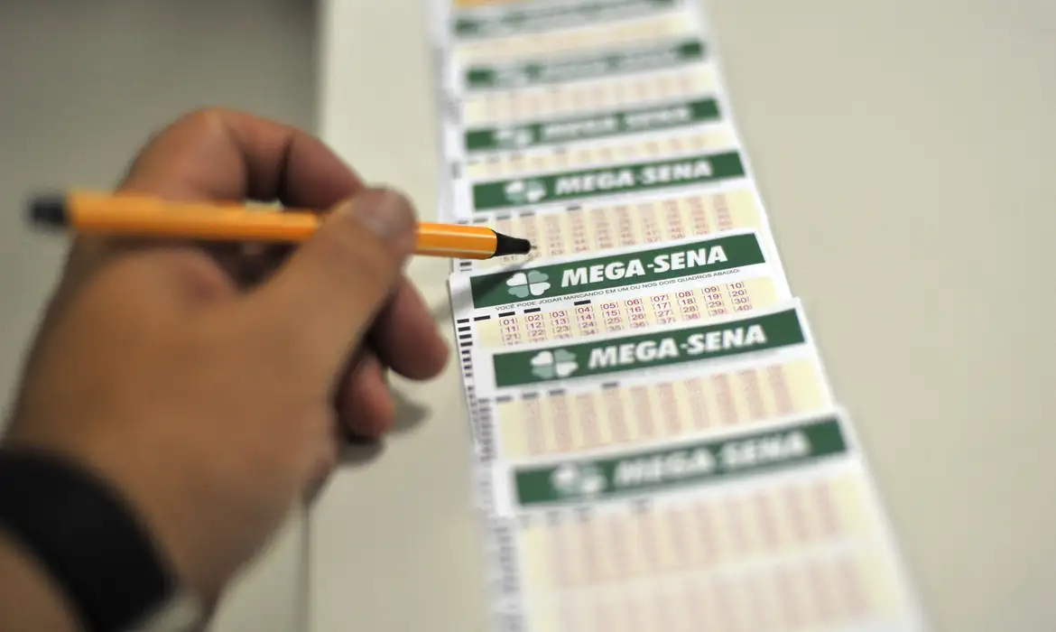 Mega-Sena não tem ganhadores e prêmio sobe para R$ 65 milhões 2 Mão masculina segurando uma caneta laranja e preta enquanto marca números em um volante da Mega-Sena. Outros cartões de aposta estão alinhados em sequência sobre uma mesa branca.