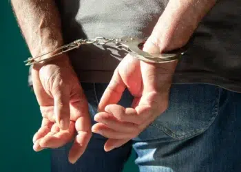 Homem algemado com as mãos para trás vestindo camiseta cinza e jeans
