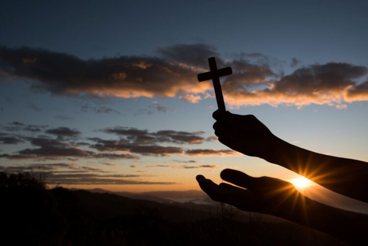 Silhueta de mãos segurando uma pequena cruz de madeira contra o céu do amanhecer em tons de laranja e azul, com o sol surgindo entre montanhas.