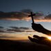 Silhueta de mãos segurando uma pequena cruz de madeira contra o céu do amanhecer em tons de laranja e azul, com o sol surgindo entre montanhas.