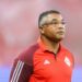 Técnico de camisa vermelha observa jogo à beira do campo com semblante sério