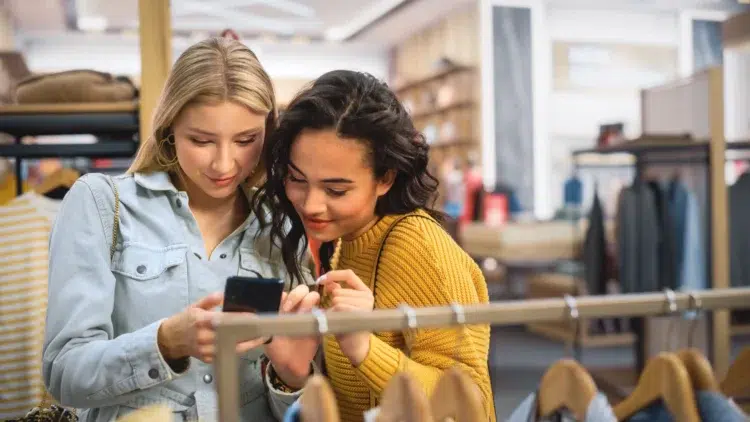 Descontos da Semana do Consumidor chegam a shoppings, bancos e e-commerces; VEJA! 2 Duas mulheres em uma loja de roupas olhando o celular enquanto conferem ofertas da Semana do Consumidor.
