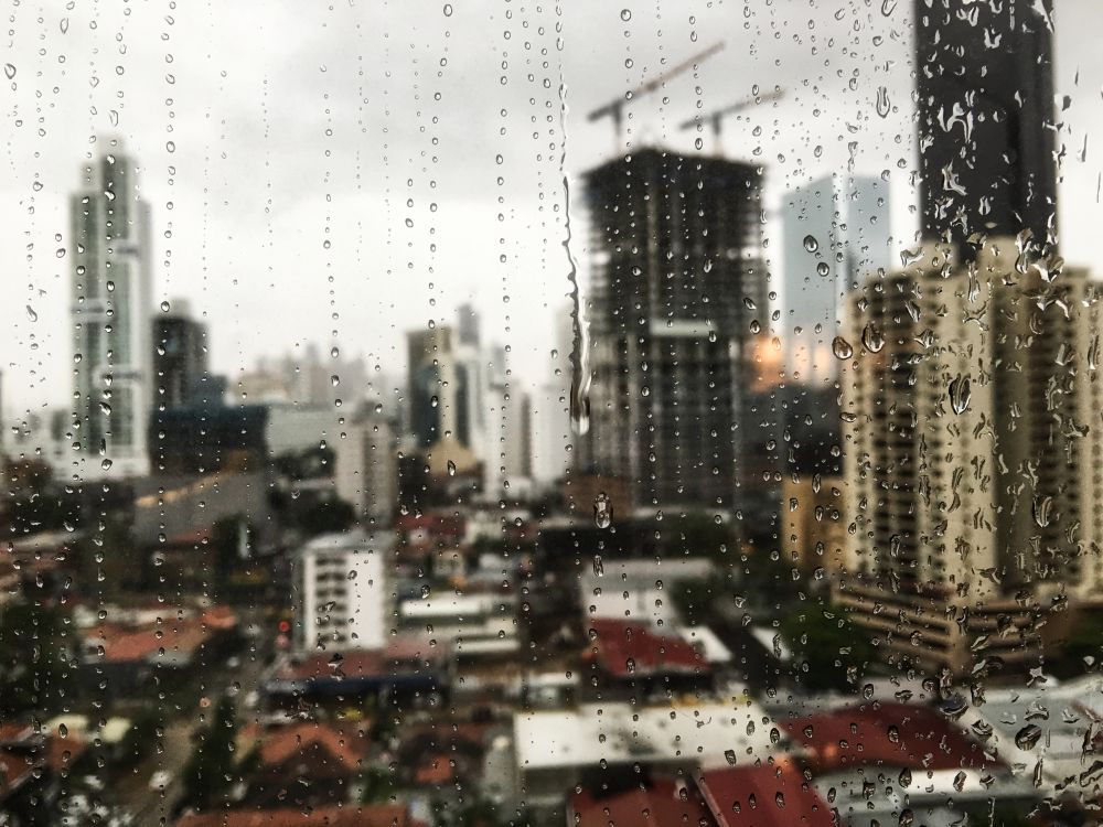 Fim de semana terá temporais, chuva forte e muito calor; confira a previsão 2 Close de um vidro de janela molhado por gotas de chuva escorrendo verticalmente. Pelo vidro embaçado, vê-se a paisagem urbana de uma cidade desfocada com diversos arranha-céus cinzas e telhados vermelhos, sob um céu nublado e opaco. No fundo, duas gruas de construção em destaque.