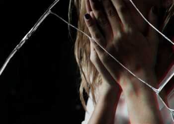 Uma mulher com as mãos cobrindo o rosto em sinal de angústia, vista através de um vidro quebrado, simbolizando a violência de gênero.