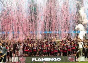 Jogadores do Flamengo reunidos no pódio, levantando a taça de campeão sob uma chuva de papéis picados cor-de-rosa e fogos de artifício ao fundo.