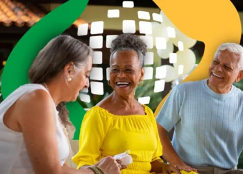 Três idosos sorridentes em ambiente externo: mulher de branco, mulher de amarelo com brincos e homem de blusa cinza