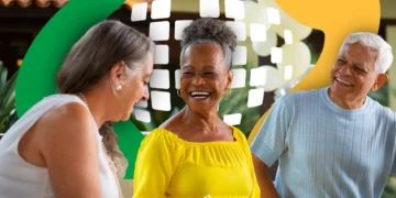 Três idosos sorridentes em ambiente externo: mulher de branco, mulher de amarelo com brincos e homem de blusa cinza