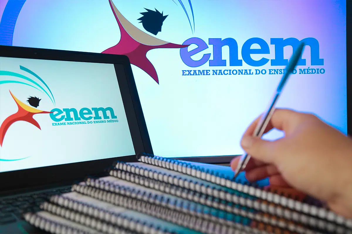 Isenção da taxa do Enem 2026 deve ser solicitada até 24 de abril 2 Estudante escreve em caderno com caneta enquanto notebook exibe logo do Enem ao lado de tela maior com marca do exame