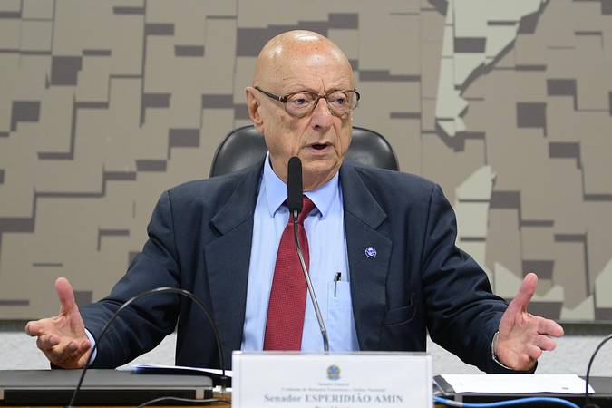 Placas de veículos no Brasil podem mudar; veja o que propõe o projeto 2 Rosto e tronco do Senador Esperidião Amin, careca e de óculos, vestindo terno azul escuro e gravata vermelha. Ele está sentado em uma mesa de debates, gesticulando com as duas mãos abertas, em frente a um microfone e uma placa de identificação com seu nome.