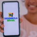 Mulher sorrindo mostra celular com a tela exibindo o logo Desenrola Brasil em fundo branco