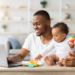 Homem sorrindo usa laptop enquanto segura bebê com brinquedo colorido em mesa clara de sala iluminada