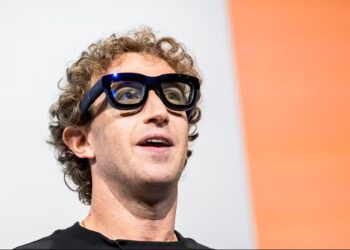 Mark Zuckerberg usando óculos inteligentes pretos grandes, fundo branco com faixa laranja, expressão atenta