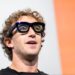 Mark Zuckerberg usando óculos inteligentes pretos grandes, fundo branco com faixa laranja, expressão atenta