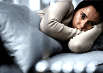 Mulher de cabelos escuros deitada em sofá cinza, com expressão triste, apoiando a cabeça nos braços