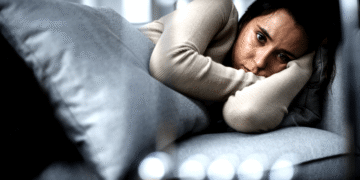 Mulher de cabelos escuros deitada em sofá cinza, com expressão triste, apoiando a cabeça nos braços